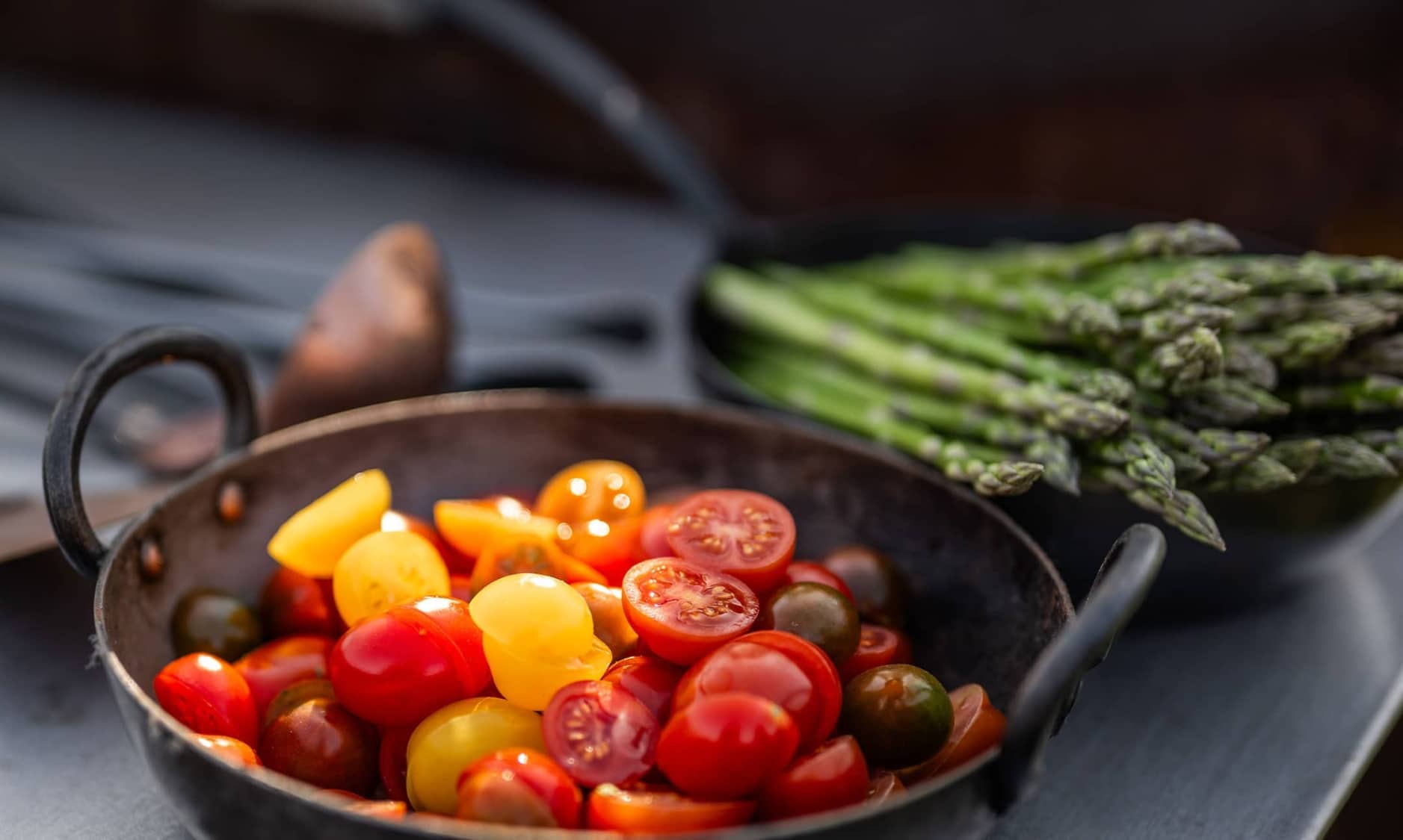 Freshly chopped cherry tomatoes in a rustic pan, with a bundle of asparagus in the background ready for an outdoor dining experience.