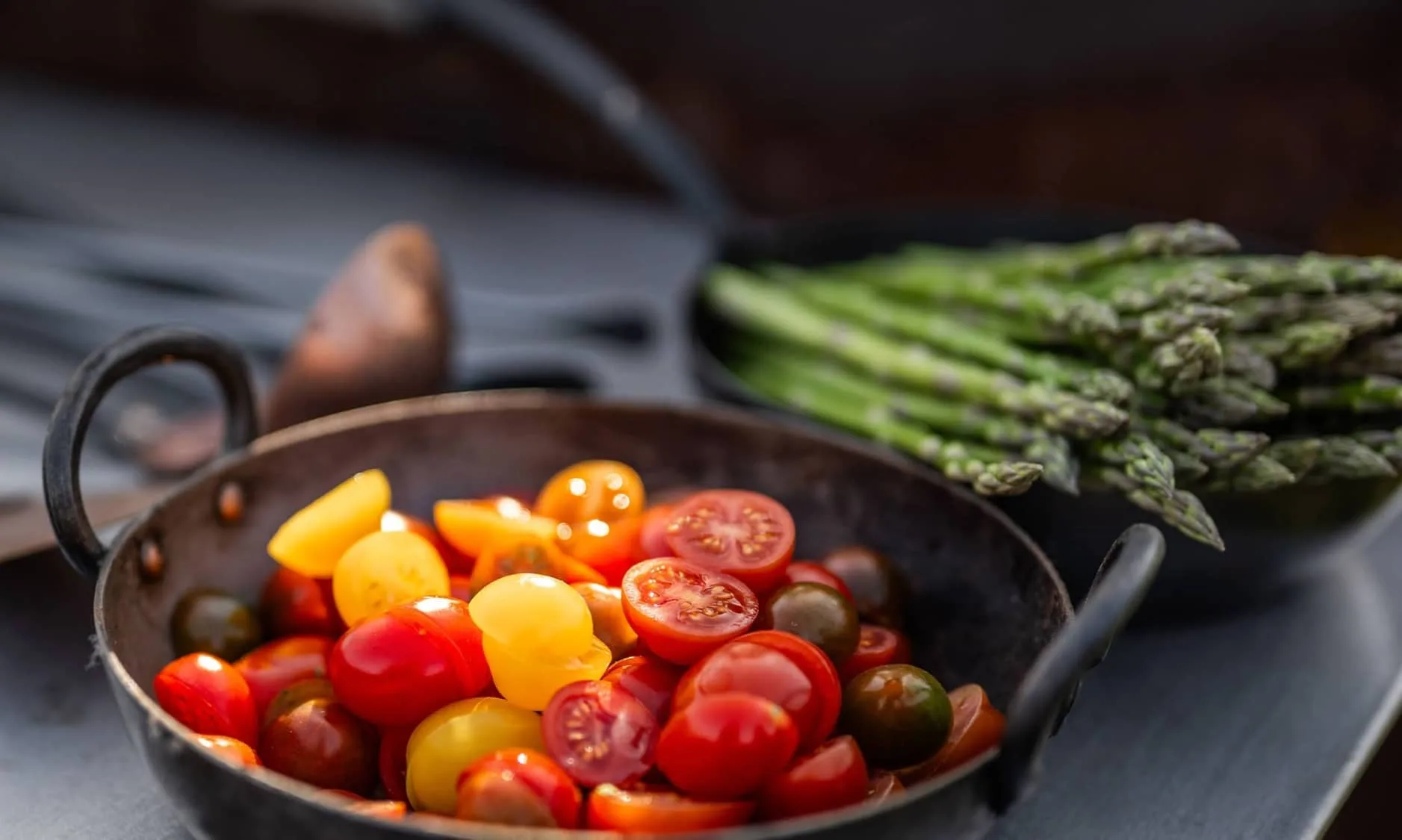 Freshly chopped cherry tomatoes in a rustic pan, with a bundle of asparagus in the background ready for an outdoor dining experience.