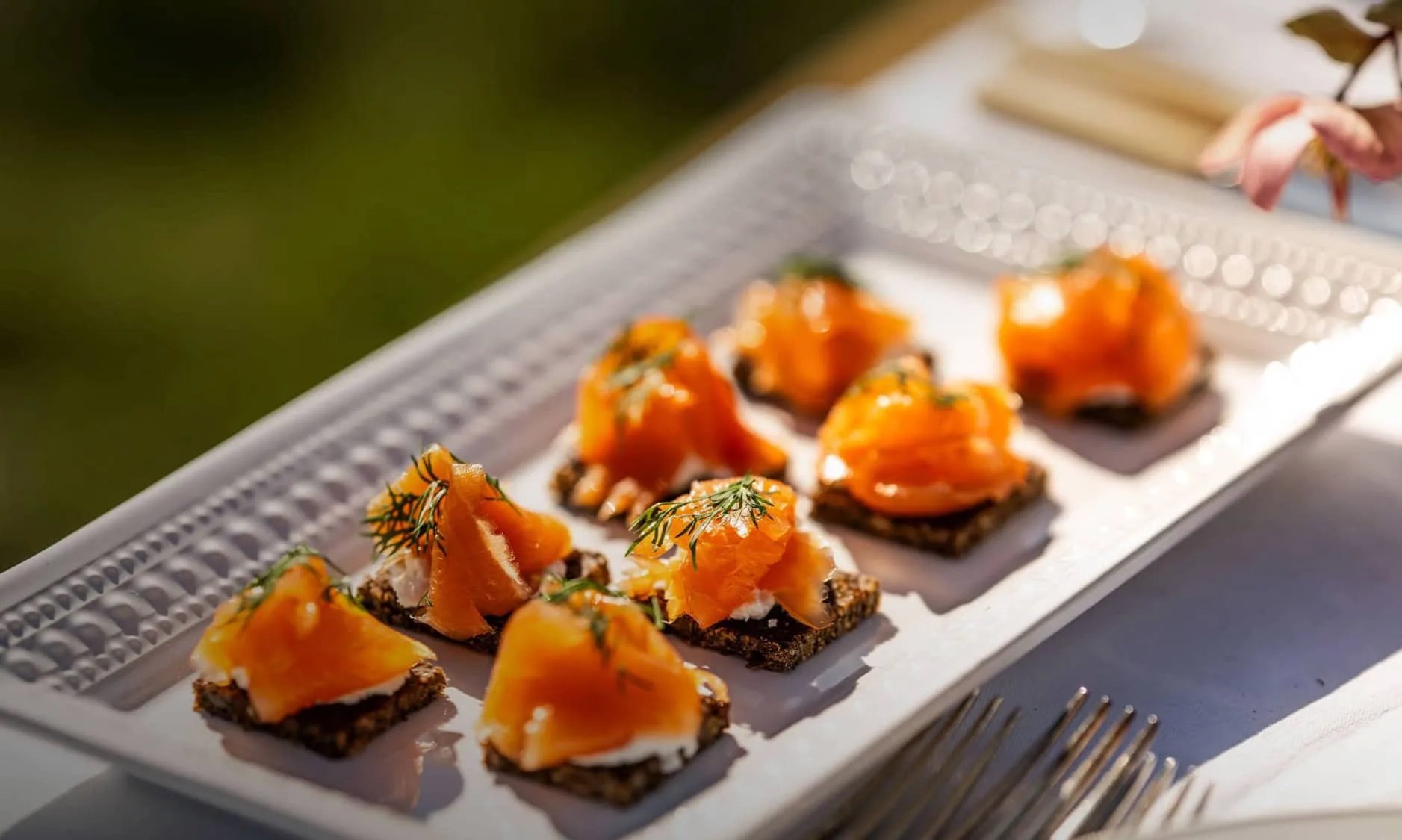 Close-up of smoked salmon canapés on rye bread, garnished with dill and served on a white platter during an elegant outdoor dining experience.